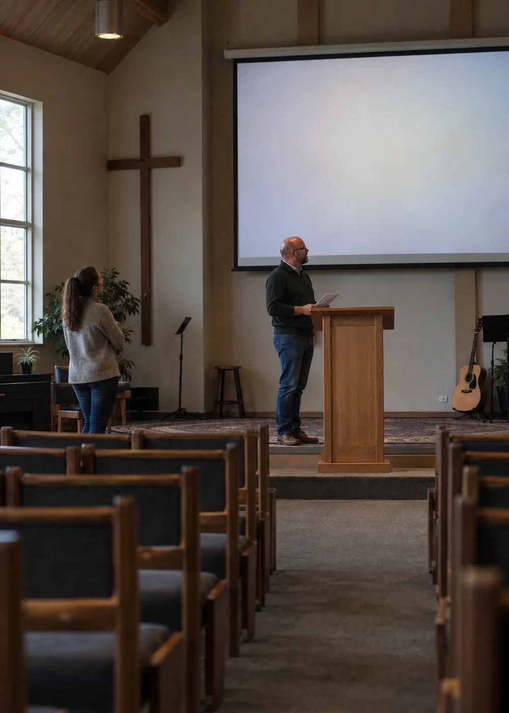 A pastor stands at a lectern with printed notes while a volunteer checks the blank projection screen at the front of a church sanctuary.